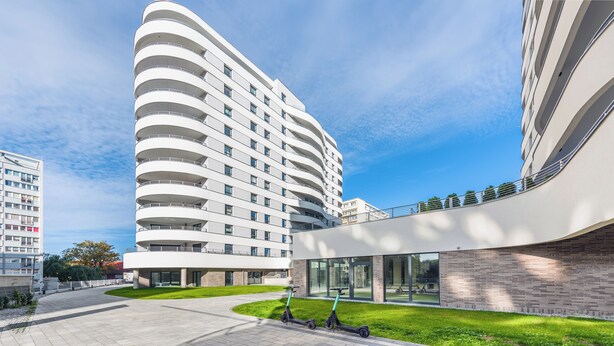 A modern building with curved balconies and a flat roof, surrounded by a well-maintained lawn and a paved walkway.