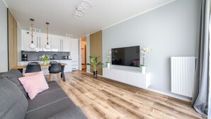 A modern living room with a sofa, a dining area with a table and chairs, a kitchen with white cabinets, and a TV mounted on the wall.