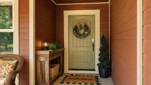 Una puerta de madera con un adorno de guirnalda, un felpudo, un farol, una silla de mimbre y una planta en maceta.