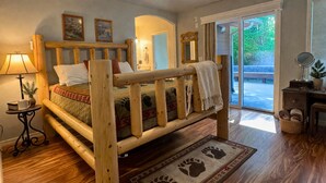 Una cama de madera con cabecera, una mesita de noche con lámpara, una planta pequeña y una ventana con vistas a vegetación.