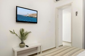 Quarto com cama, televisão na parede, mesa de cabeceira branca pequena com uma planta e uma porta.