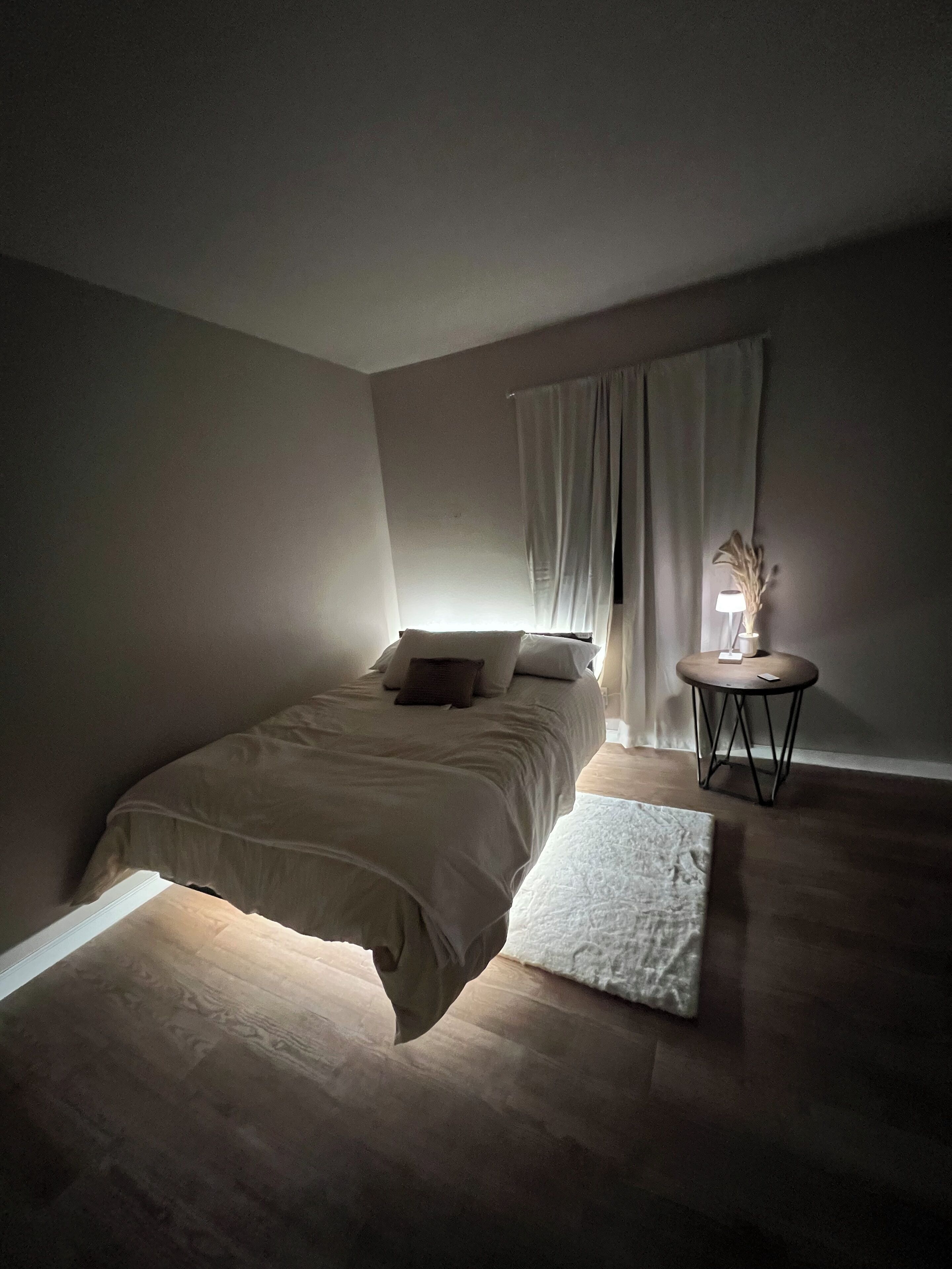 Une chambre à coucher avec un lit, une petite table ronde avec une lampe et une fenêtre avec des rideaux.