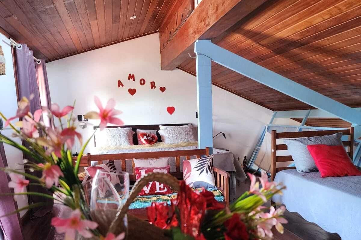 Een gezellige kamer met een houten plafond, een bed met rode en witte beddengoed, een muur met 'AMOR' en hartversieringen, en een bloemenmand op de vloer.
