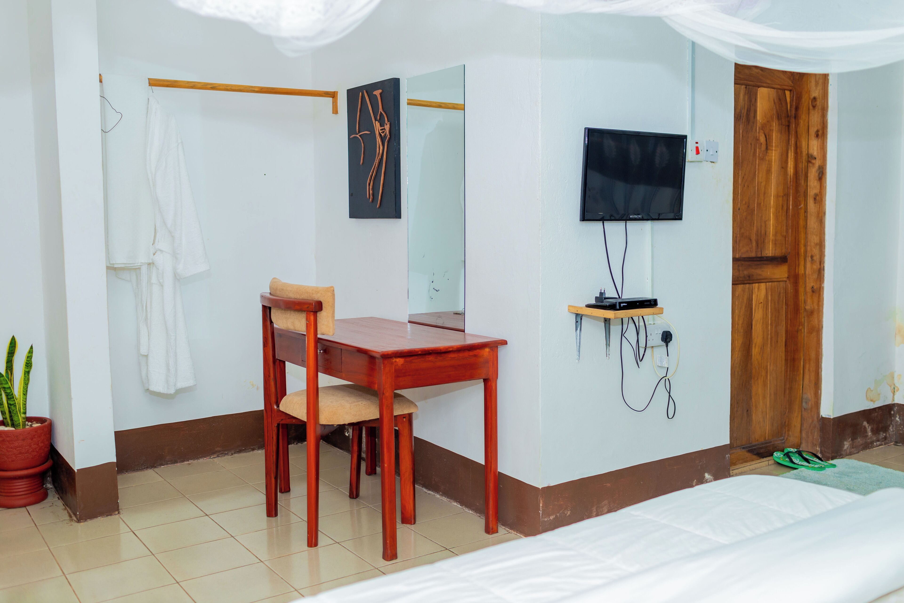 Ein Hotelzimmer mit Bett, einem Holztisch mit Stuhl, einem an der Wand befestigten Fernseher, einer Holztür und einer Topfpflanze.