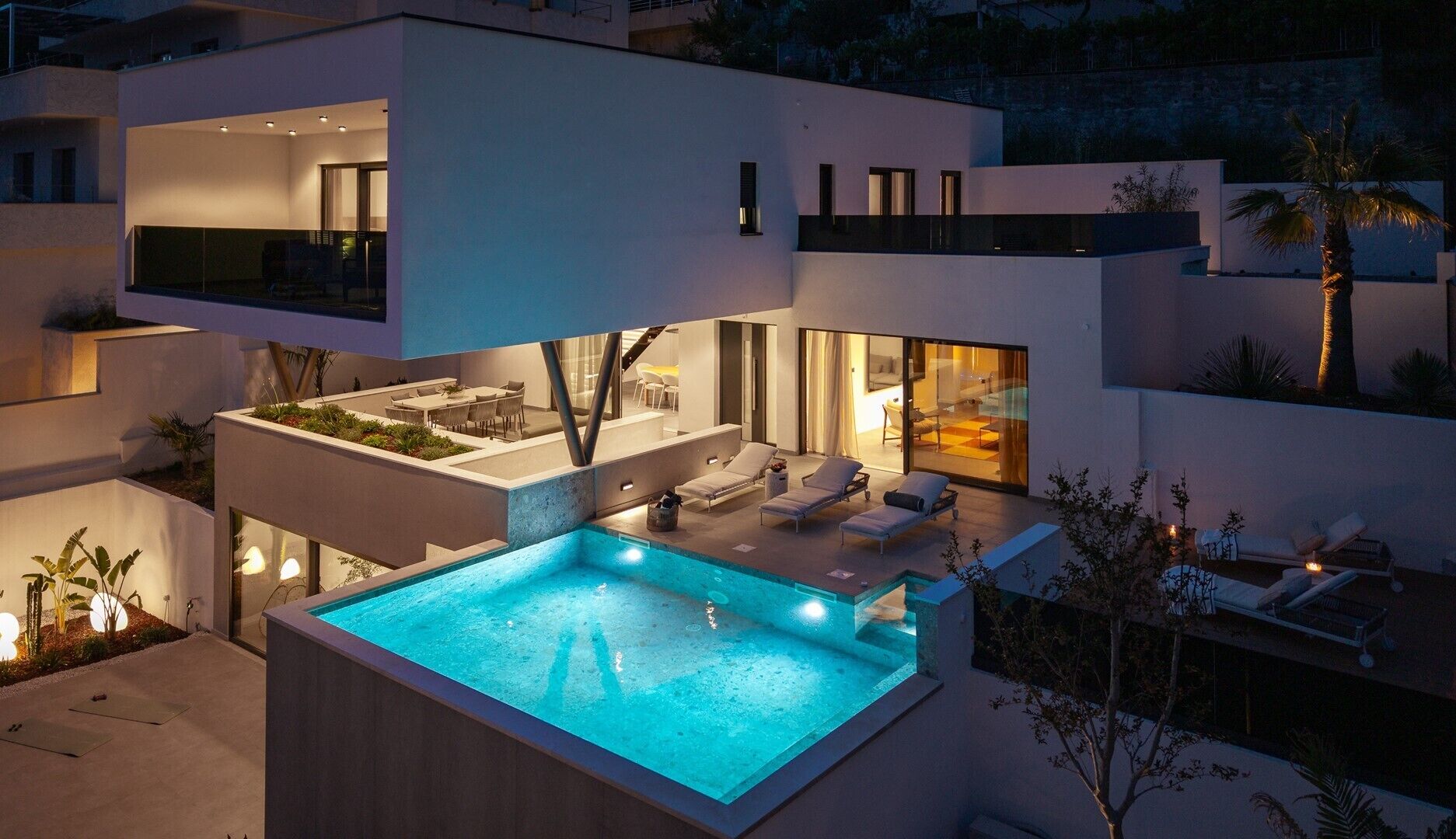 A modern multi-level house with a rooftop pool, outdoor seating, and a well-lit interior.