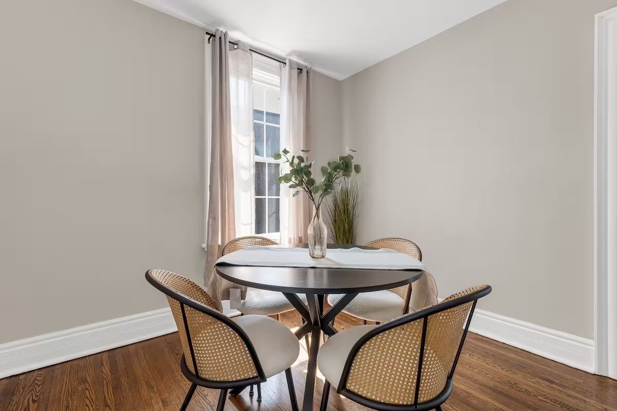 Un coin repas avec une table ronde, quatre chaises en osier, un vase avec des plantes vertes et une fenêtre avec des rideaux.