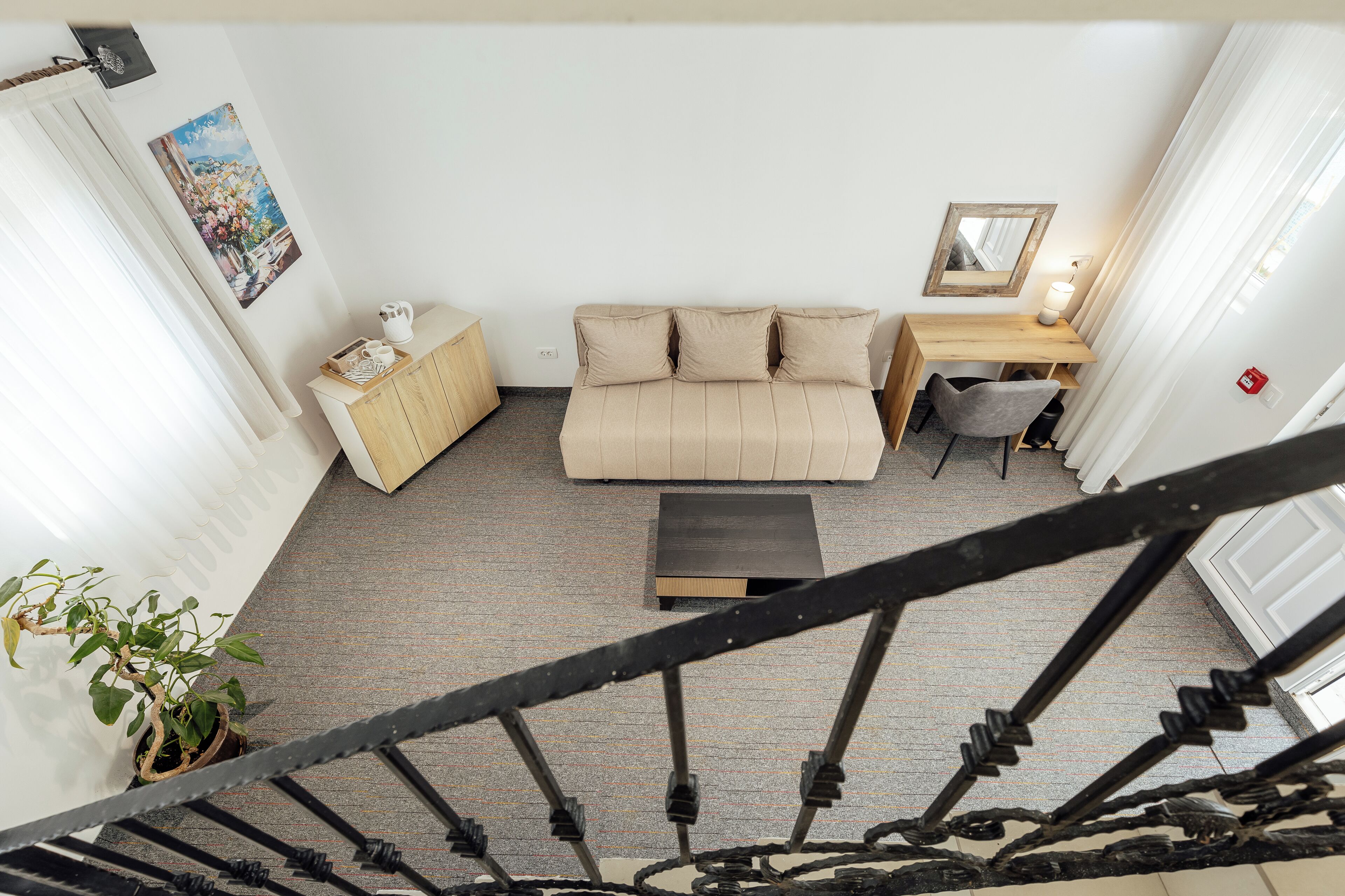 A room with a sofa, a desk, a chair, a small table, a potted plant, and a staircase.