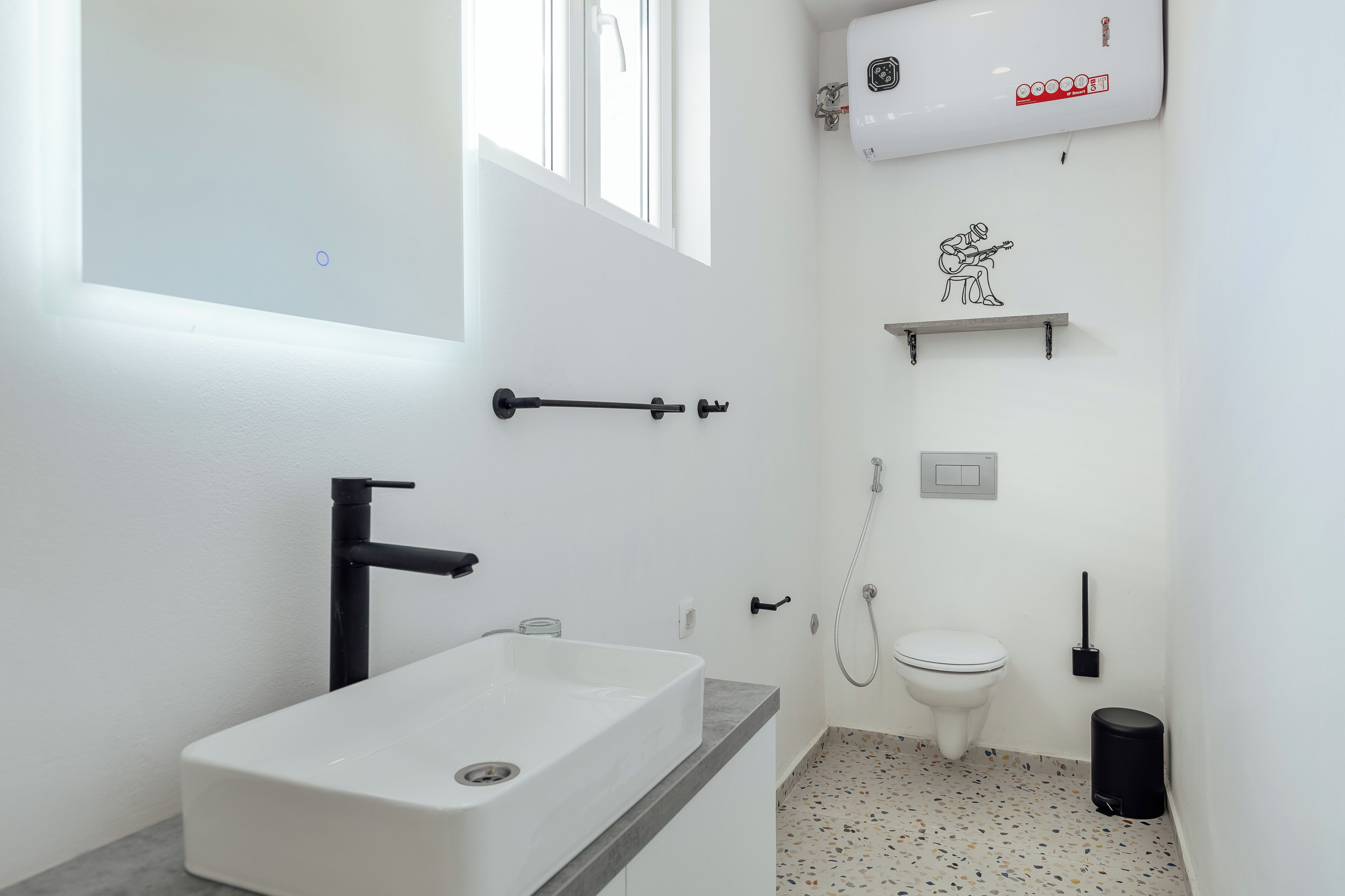 Un baño moderno con un lavamanos, inodoro y ducha blancos. Una ventana proporciona luz natural, y hay un perchero en la pared.