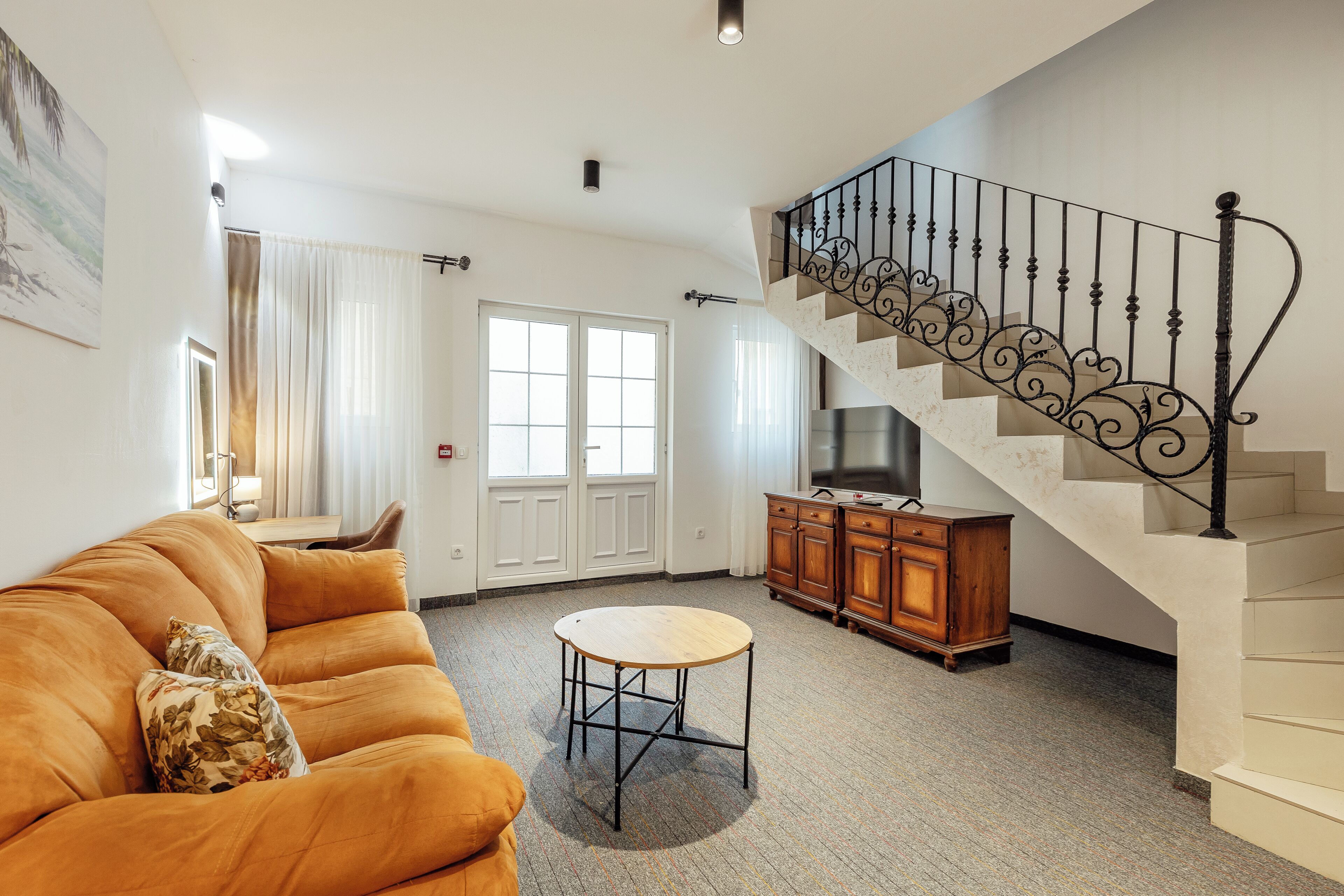 A living room with a sofa, a round table, a staircase with a decorative railing, and a TV cabinet.