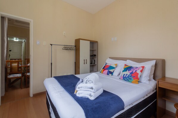 Un lit bien fait, avec une couverture bleue et des oreillers blancs, une armoire en bois et une chaise dans une pièce aux murs de couleur beige.