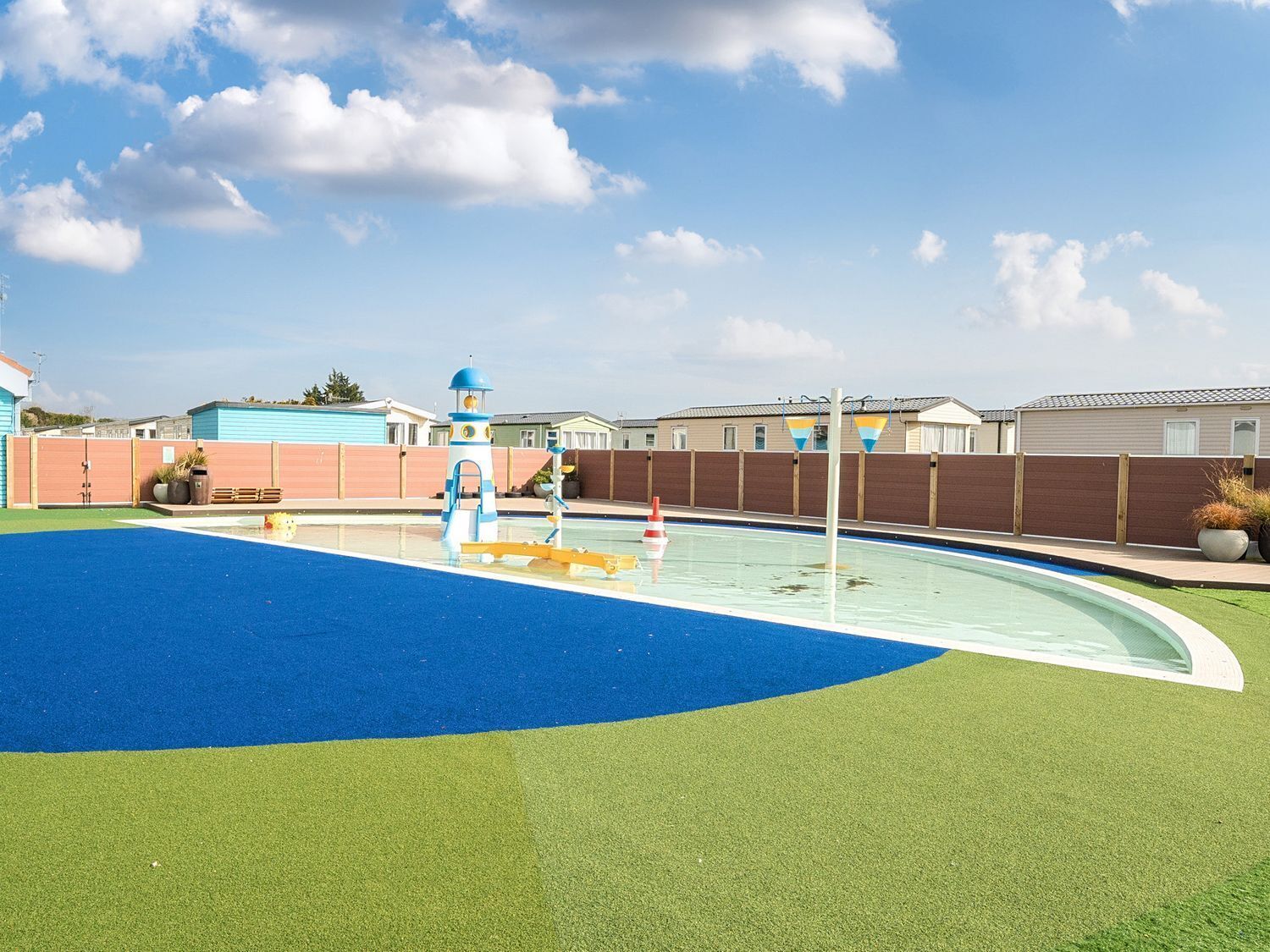 Una piscina con superficie azul y verde, rodeada por una valla de madera y casas al fondo.
