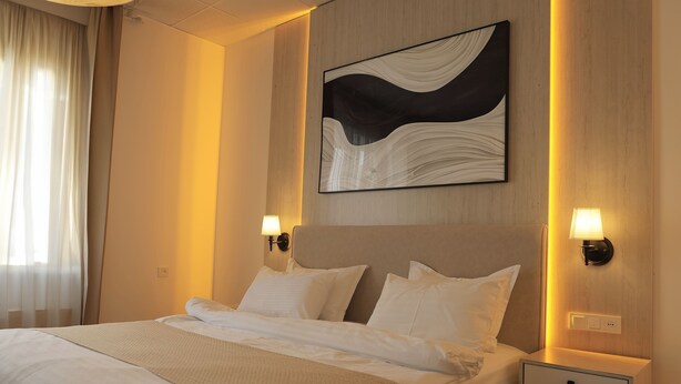 A hotel room with a large bed, two bedside tables with lamps, a wall-mounted abstract painting, and a window with sheer curtains. - UNO Hotel Jalal-Abad (Jalal-Abad)
