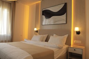 Habitación de hotel con una cama grande, dos mesitas de noche con lámparas, un cuadro abstracto en la pared y una ventana con cortinas traslúcidas.