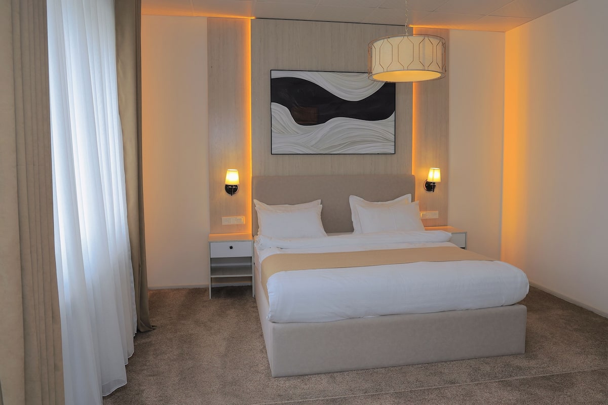 A hotel room with a large bed, two bedside tables, a wall-mounted artwork, and two wall-mounted lamps.