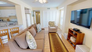 A living room with a beige sofa, a chair, a TV, and a kitchen area in the background.