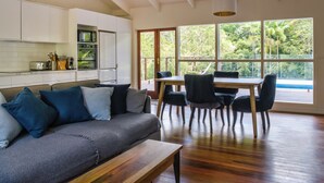 A modern kitchen with white cabinets, a dining area with a wooden table and chairs, and large windows offering a view of greenery.