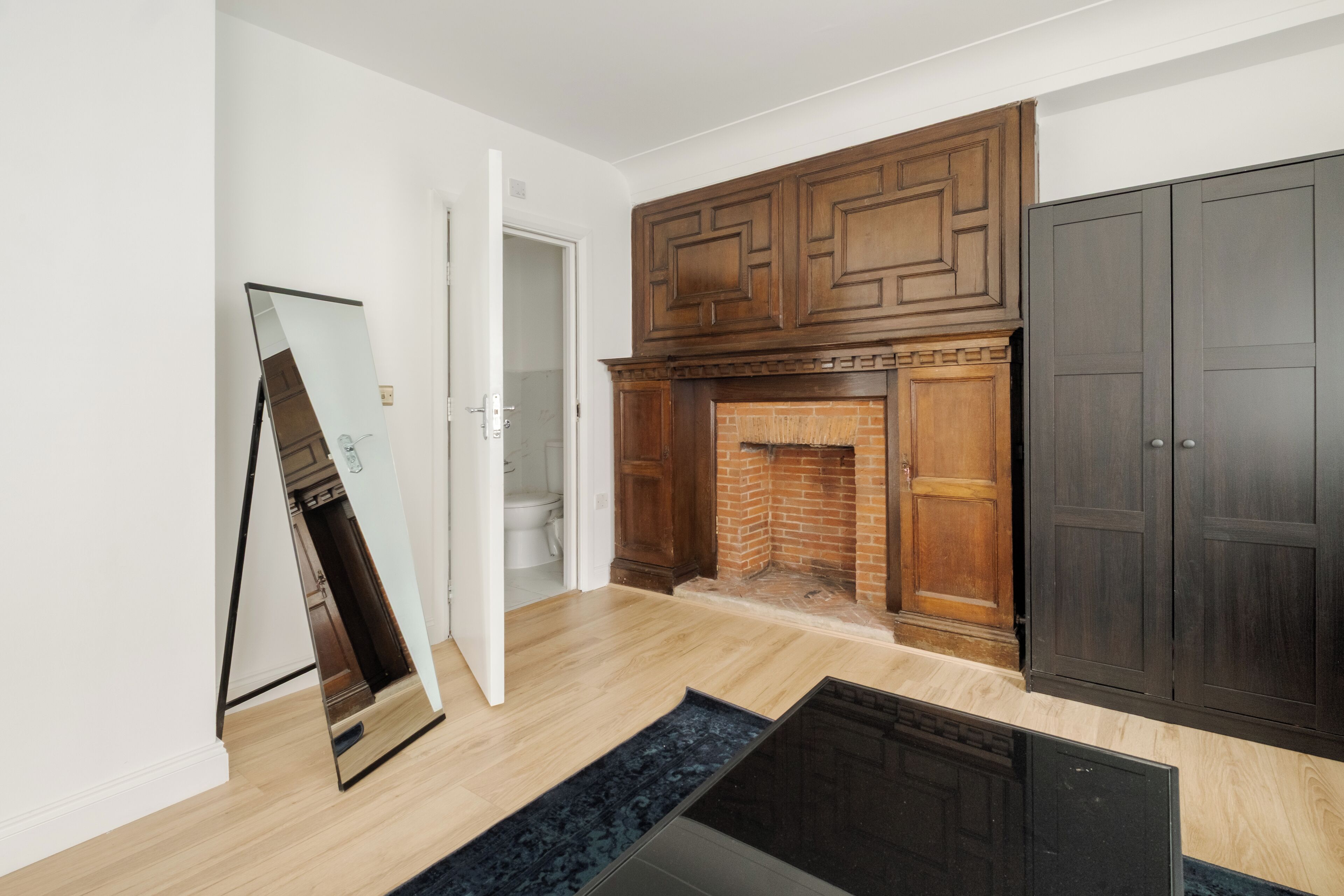 A room with a wooden fireplace, a large mirror, and a dark wardrobe.