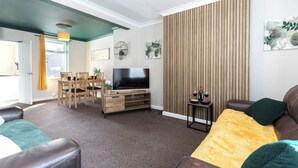 A modern living room with a flat-screen TV, a dining area with chairs and a table, and a large window with curtains.