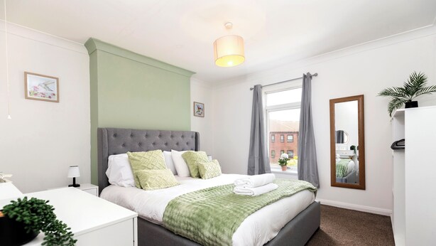 A modern bedroom with a grey upholstered bed, white bedding, a green throw blanket, a bedside table with a lamp, and a window with grey curtains.
