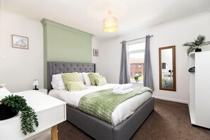 Une chambre à coucher moderne avec un lit recouvert d’un tissu gris, une literie blanche, un plaid vert, une table de chevet avec une lampe et une fenêtre ornée de rideaux gris.