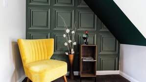 A yellow chair, a wooden side table with a vase of white flowers, and a bookshelf against a green paneled wall.