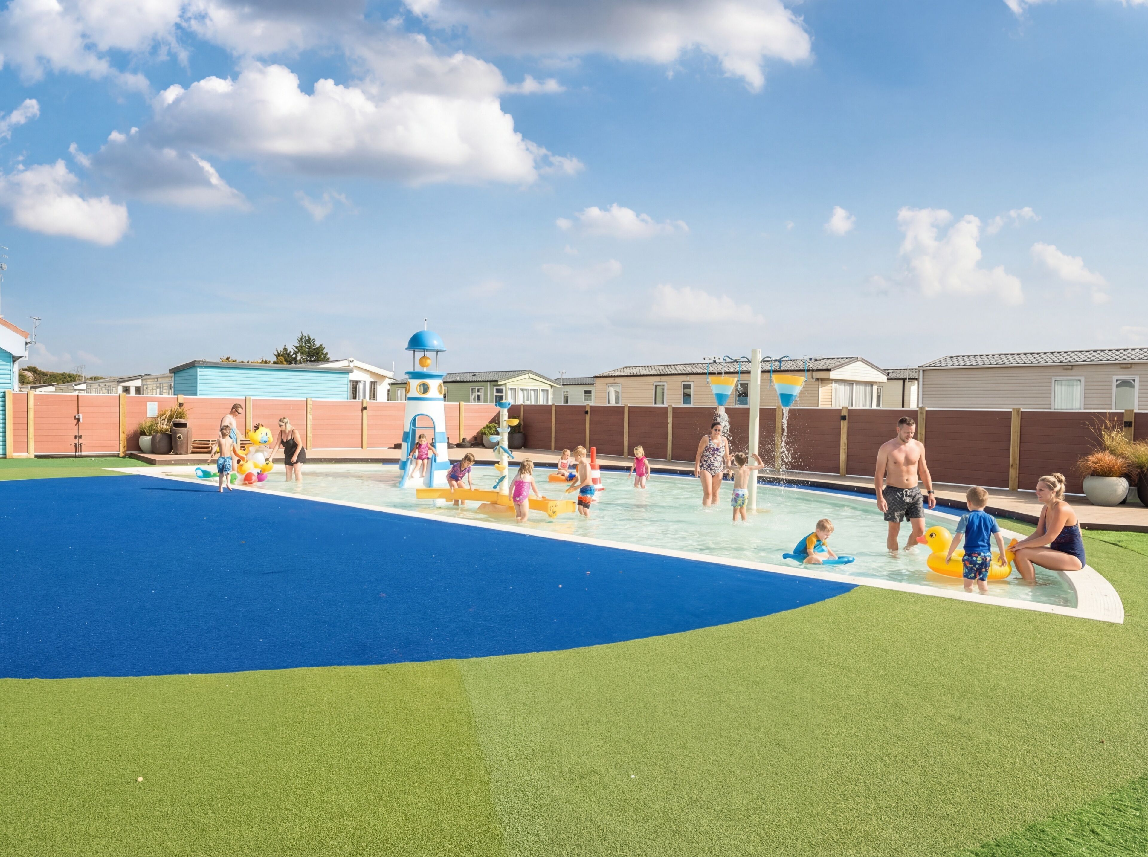 Zona de piscina con suelo de césped azul y verde, rodeada por una valla y casas al fondo.