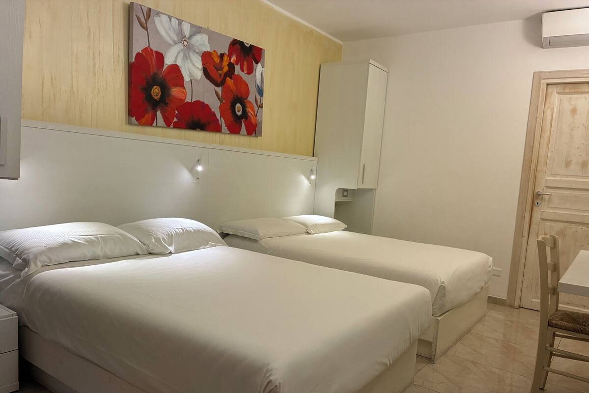 Ein Hotelzimmer mit zwei Einzelbetten, einem Holzkopfstück, einem Blumenbild, einem weißen Kleiderschrank, einem kleinen Tisch und einem Sessel.