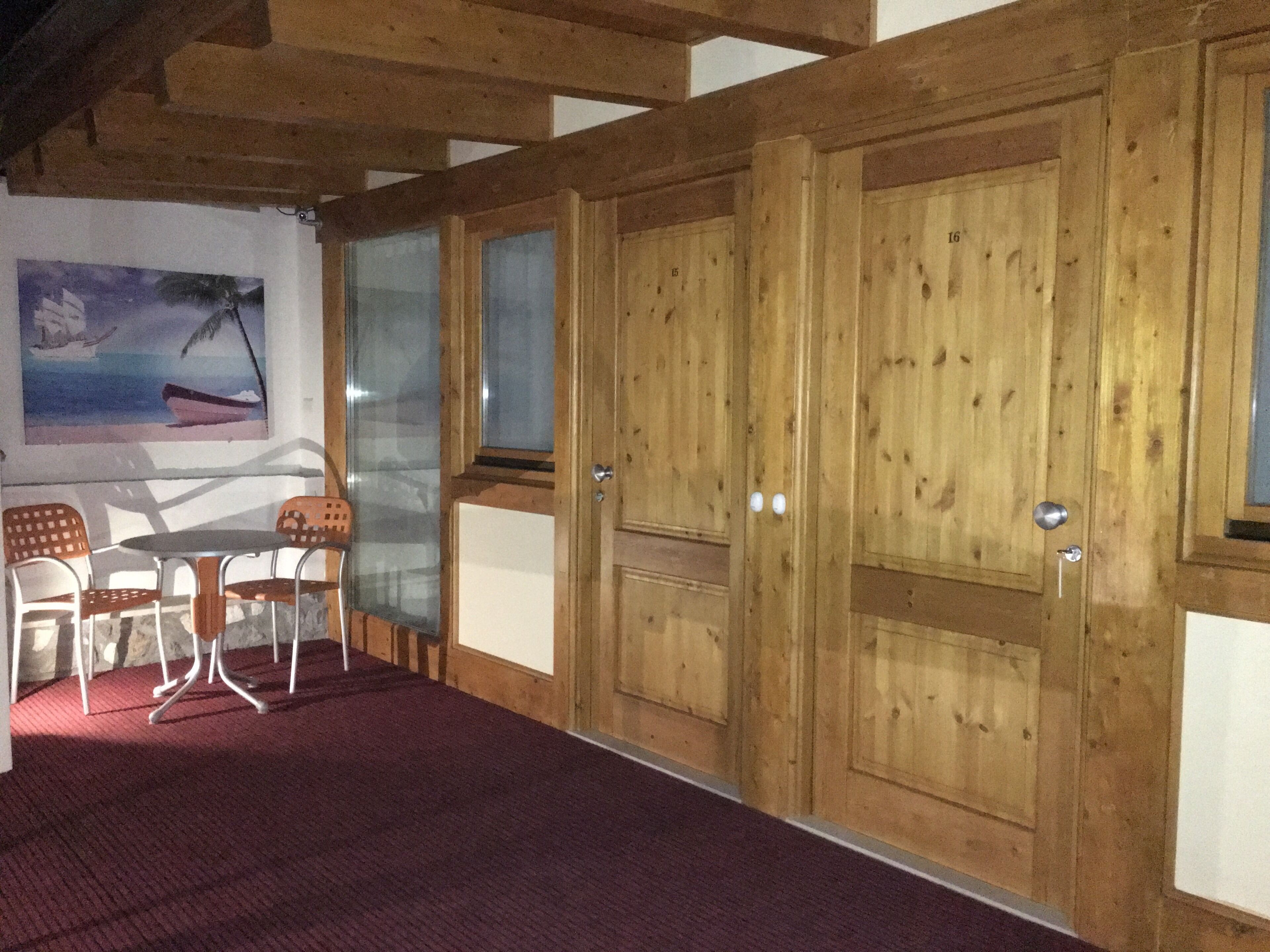 Een kamer met een houten deur, een schilderij van een boot aan de muur, een tafel met stoelen en een rood tapijt.