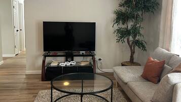 A living room with a flat-screen TV on a stand, a round coffee table, a sofa with cushions, a potted plant, and a carpeted floor.