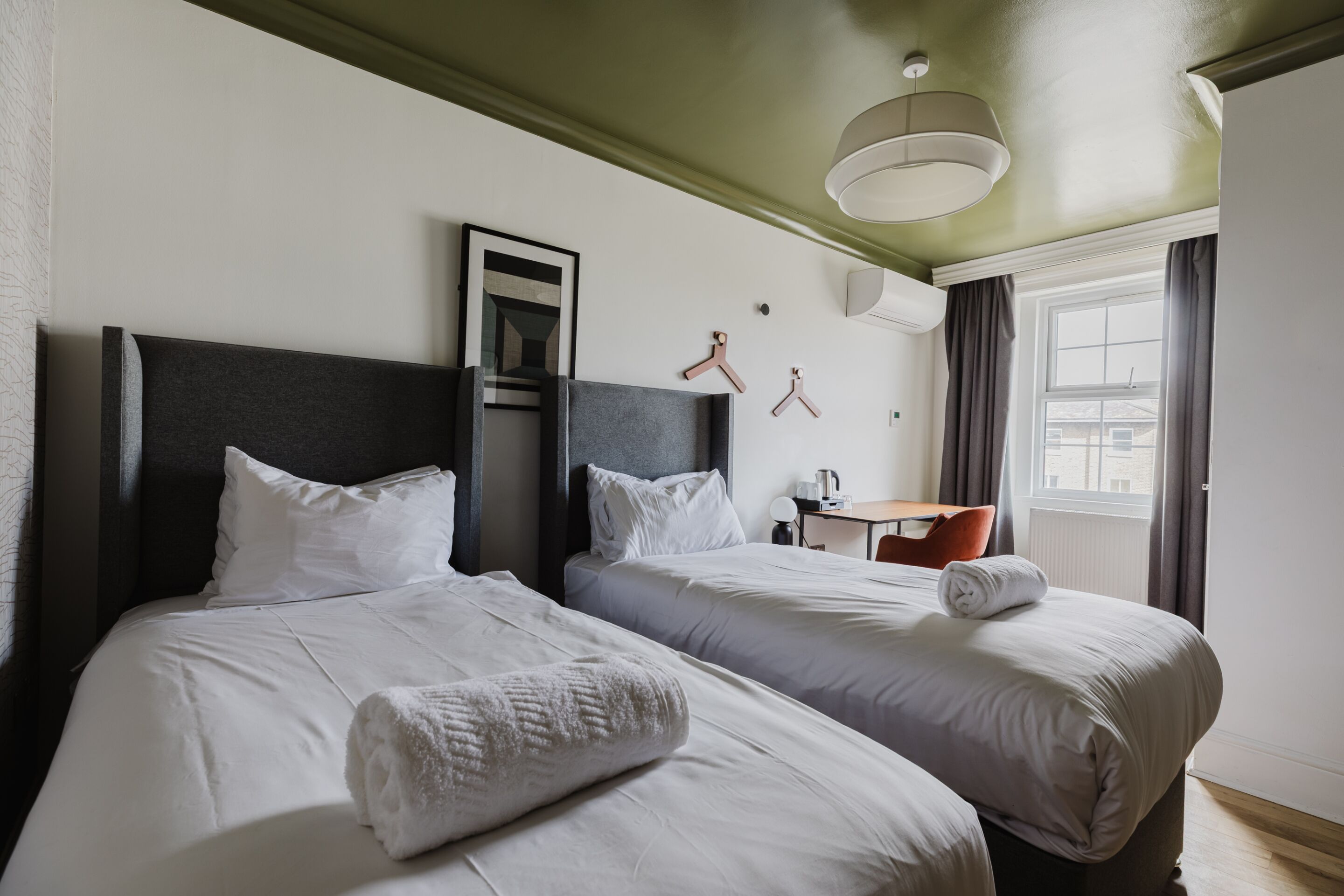 A hotel room with two beds, a desk, a chair, a window with curtains, and a hanging light fixture.