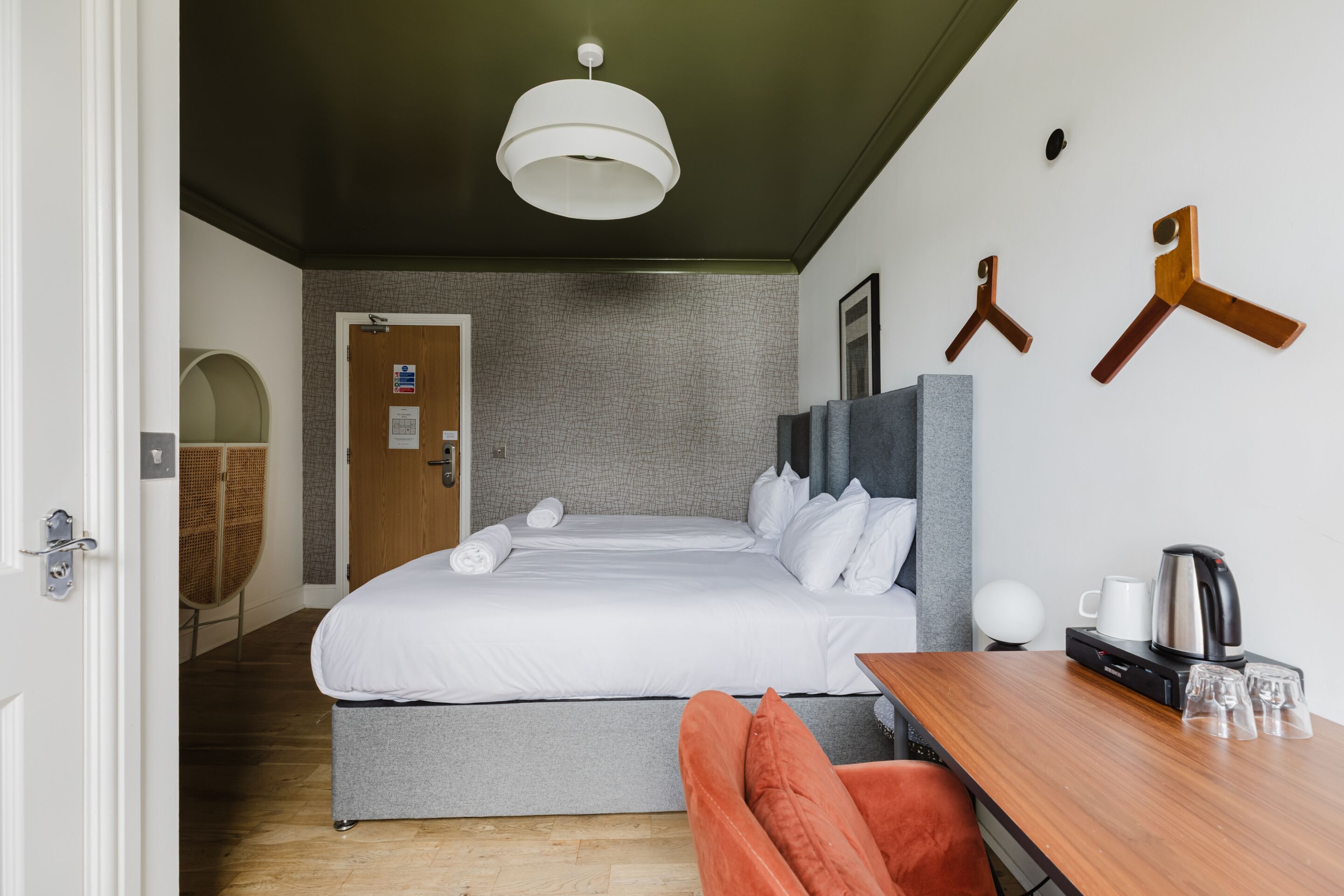 Ett hotellrum med en säng, ett träbord med en vattenkokare och glas, en hängande lampa och en dörr som leder till ett annat rum.