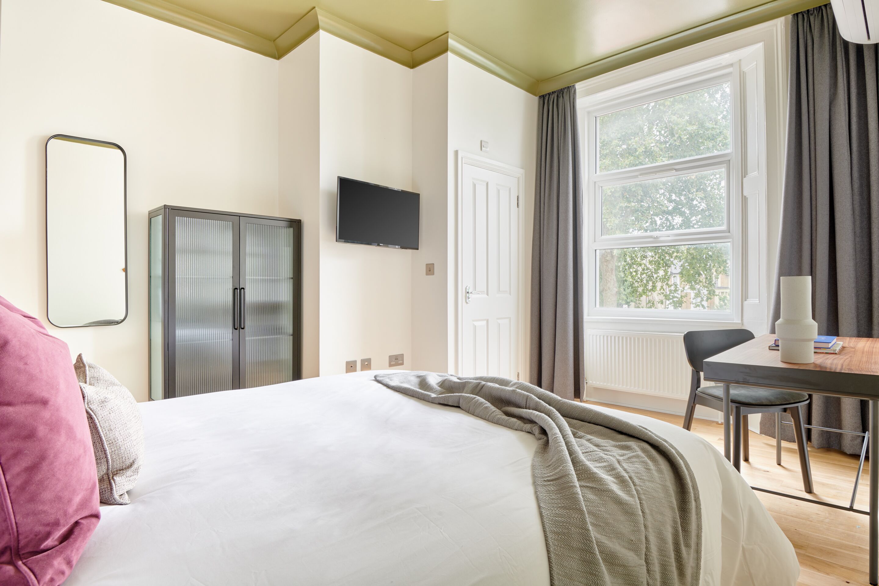 Quarto de hotel com cama, escrivaninha, televisão, guarda-roupa e janela com cortinas.