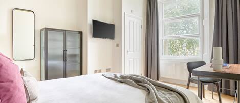 Quarto de hotel com cama, escrivaninha, televisão, guarda-roupa e janela com cortinas.