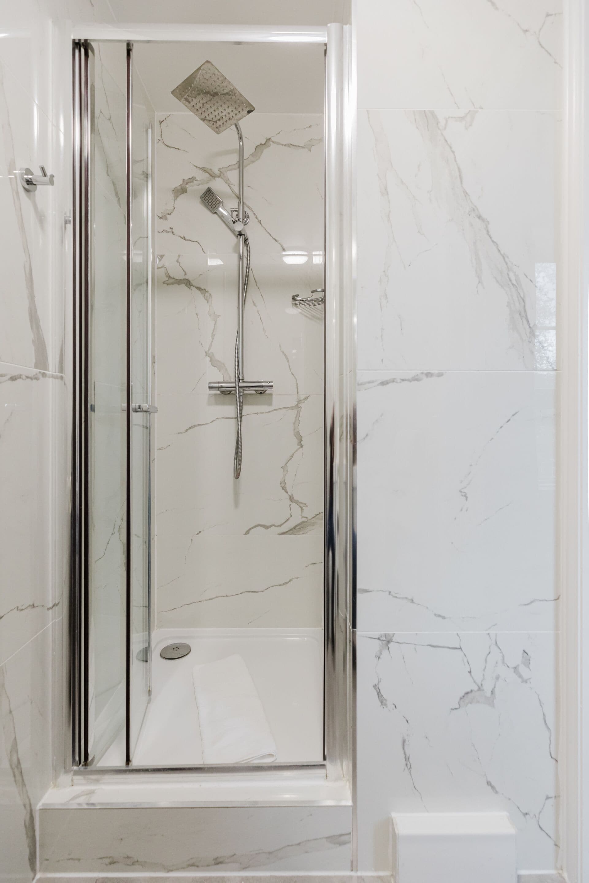 Een douche met glazen wand, voorzien van een handdouche en een douchekop, tegen een marmeren wand.