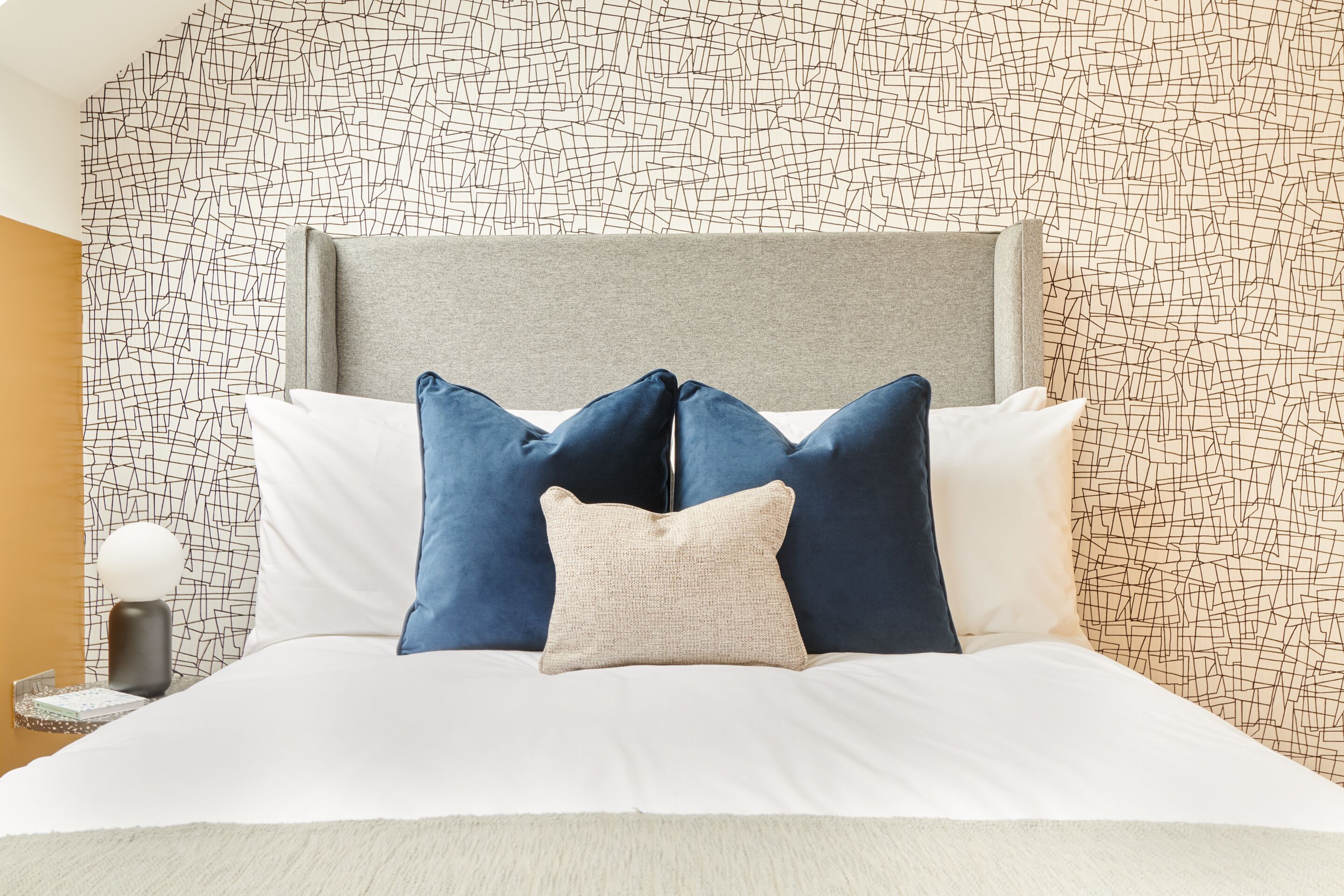 A neatly made bed with a grey headboard, white and blue pillows, and a patterned wall in the background.