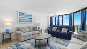 Un salon spacieux avec une grande fenêtre, un ventilateur de plafond et un tapis bleu et blanc.