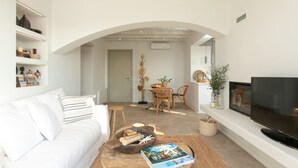 Un salon moderne avec un canapé blanc, une table basse ronde sur laquelle sont posés des magazines, un téléviseur sur un meuble et un coin repas comprenant une chaise et une table en osier.