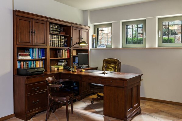 A study room with a wooden desk, bookshelves, a leather chair, and a window with a view of greenery.