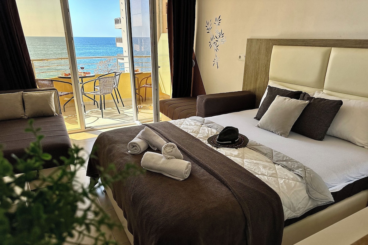 A hotel room with a bed, a chair, a table, and a view of the sea.