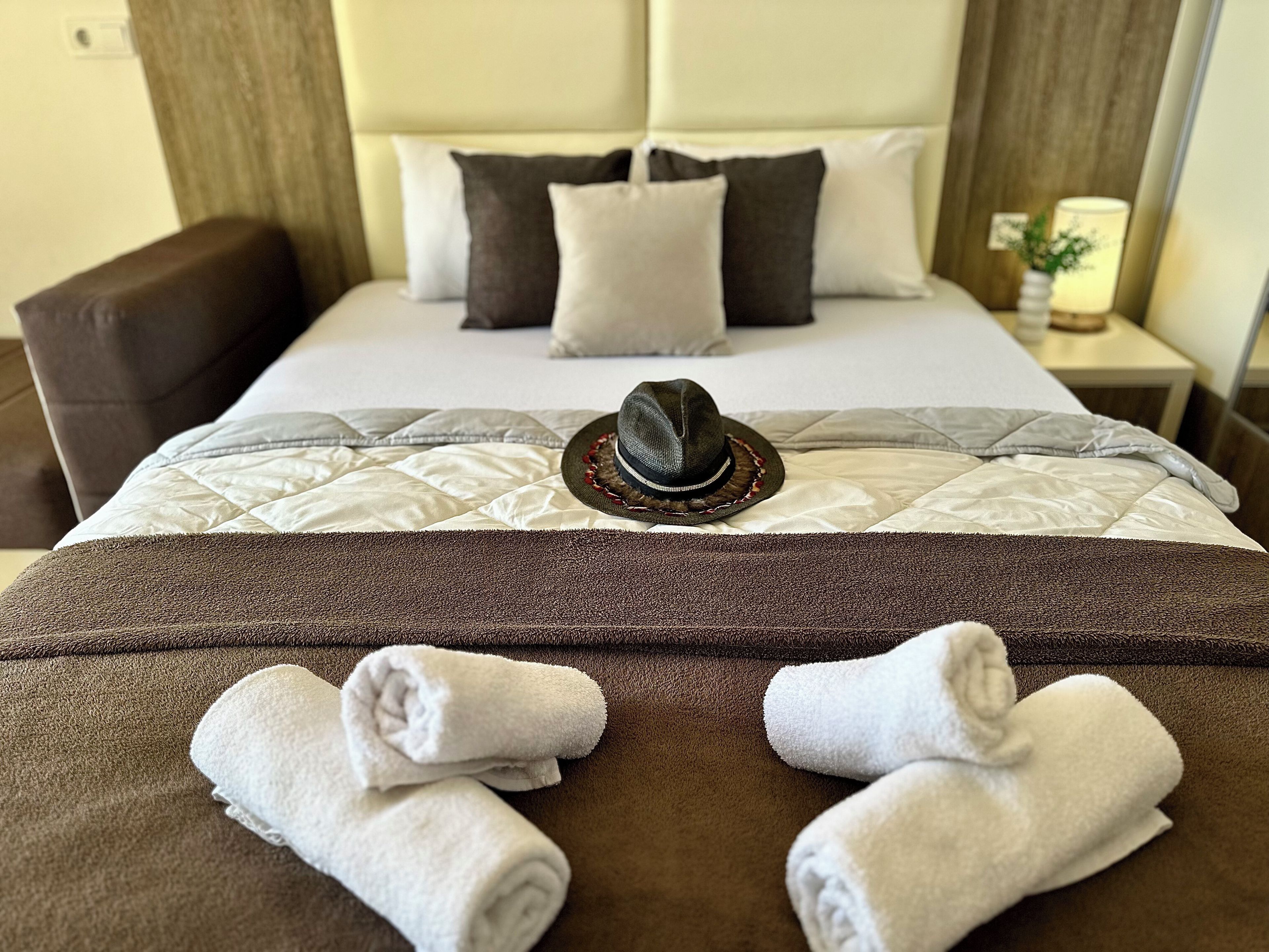 Una cama bien hecha con un sombrero y toallas, un cabecero marrón y una mesita de noche con una lámpara.