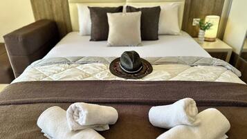 Una cama bien hecha con un sombrero y toallas, un cabecero marrón y una mesita de noche con una lámpara.