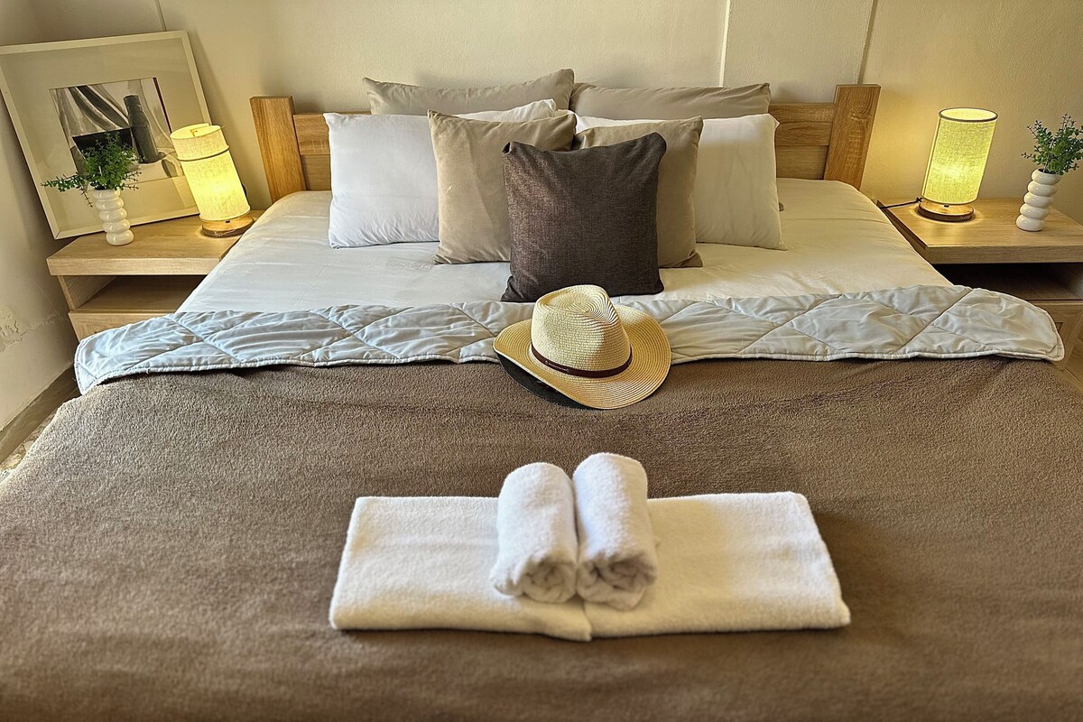 A neatly made bed with pillows, a rolled towel, and a hat on the bed.