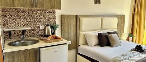 A compact hotel room with a kitchenette, a bed with pillows, and a small refrigerator.