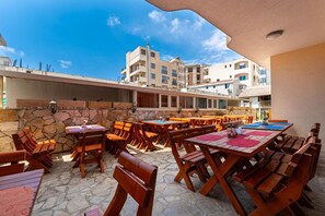 Un patio al aire libre con mesas y bancos de madera, paredes de piedra y vista a edificios residenciales en el fondo.