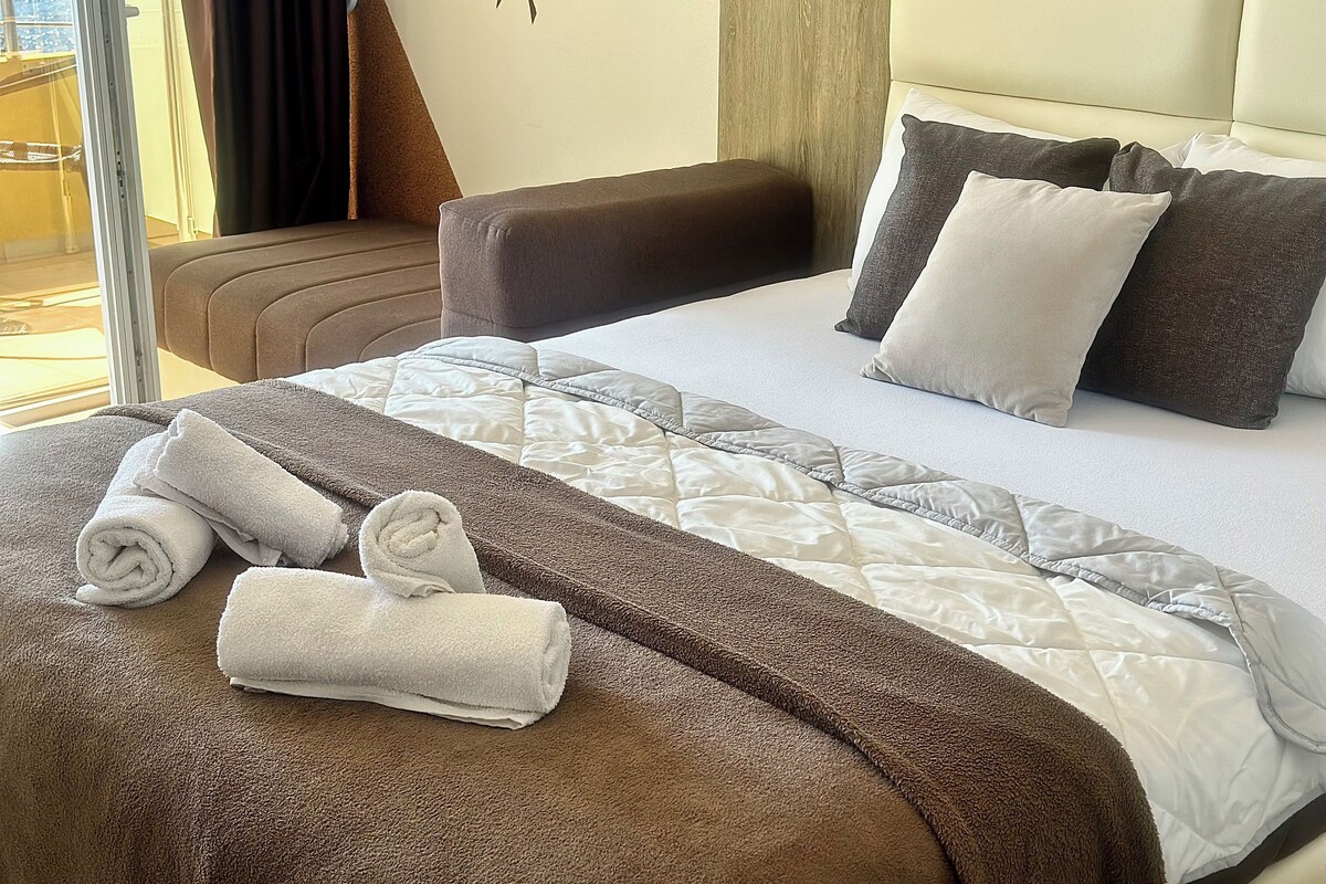 A hotel room with a bed, pillows, a brown sofa, and rolled towels.