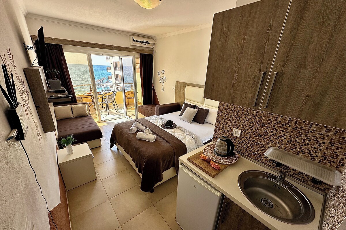 A compact hotel room with a balcony, a bed, a small kitchenette, and a view of the sea.