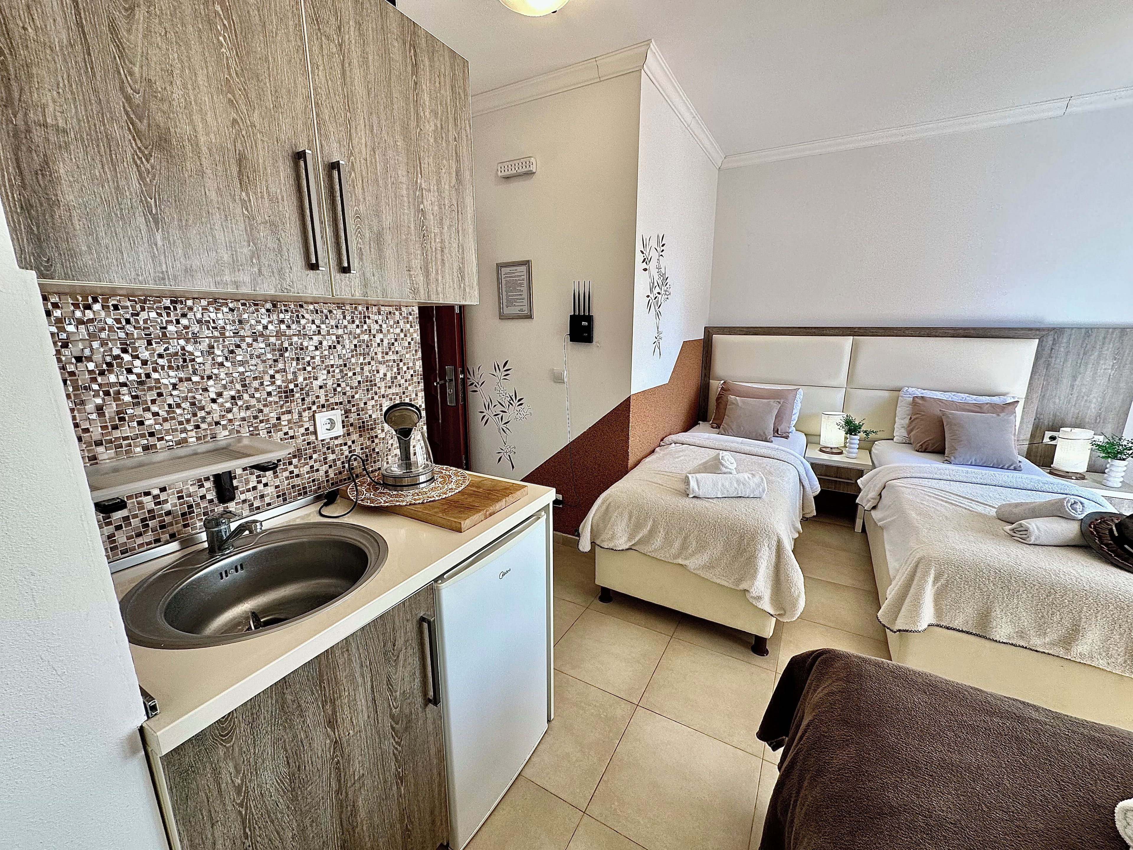 A compact hotel room with a kitchenette, a bed with two pillows, a bedside table with a lamp, and a decorative wall piece.