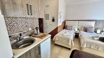 A compact hotel room with a kitchenette, a bed with two pillows, a bedside table with a lamp, and a decorative wall piece.