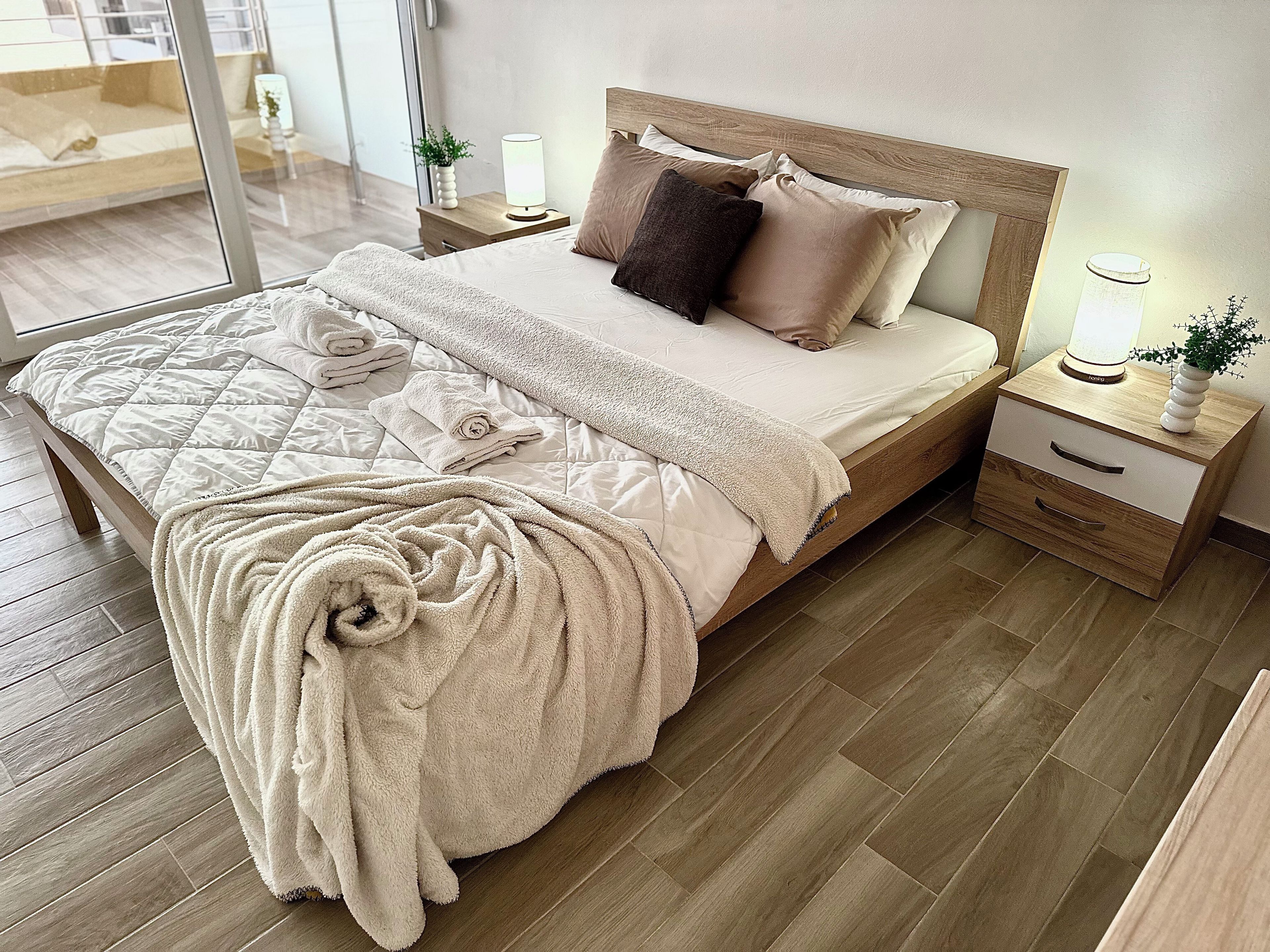 A neatly made bed with a quilt, pillows, and a blanket. There are bedside tables with lamps and a small plant. The room has wooden flooring and a large mirror on one wall.