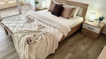 A neatly made bed with a quilt, pillows, and a blanket. There are bedside tables with lamps and a small plant. The room has wooden flooring and a large mirror on one wall.